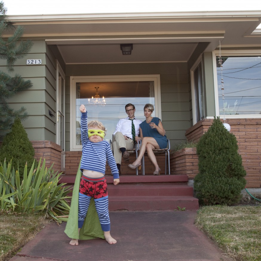 Boy in super hero costume in front of ranch-style house with parents